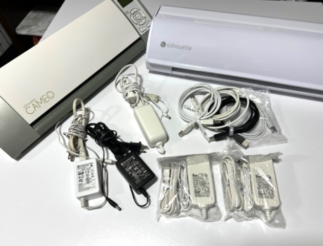 A picture of an original Silhouette Cameo and a Cameo 5 machine with several power cords and USB cables showing they all use the same cords