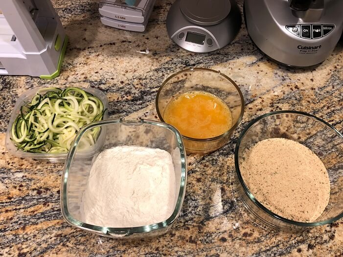 Bowls of spiral Zucchini, seasoned flour, seasoned egg wash, and seasoned bread crumbs ready for making zucchini curly fries.